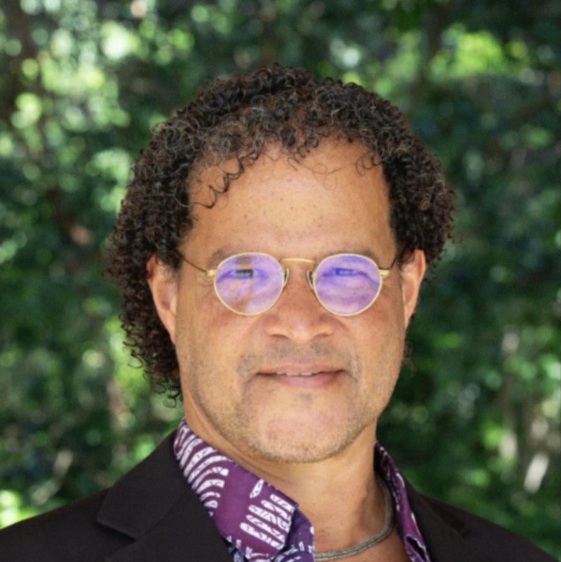 Portrait of Raphael Raphael outdoors in front of green leaves. He has curly dark hair and round gold-rimmed glasses, and wears a dark jacket over a purple patterned shirt. He is smiling.
