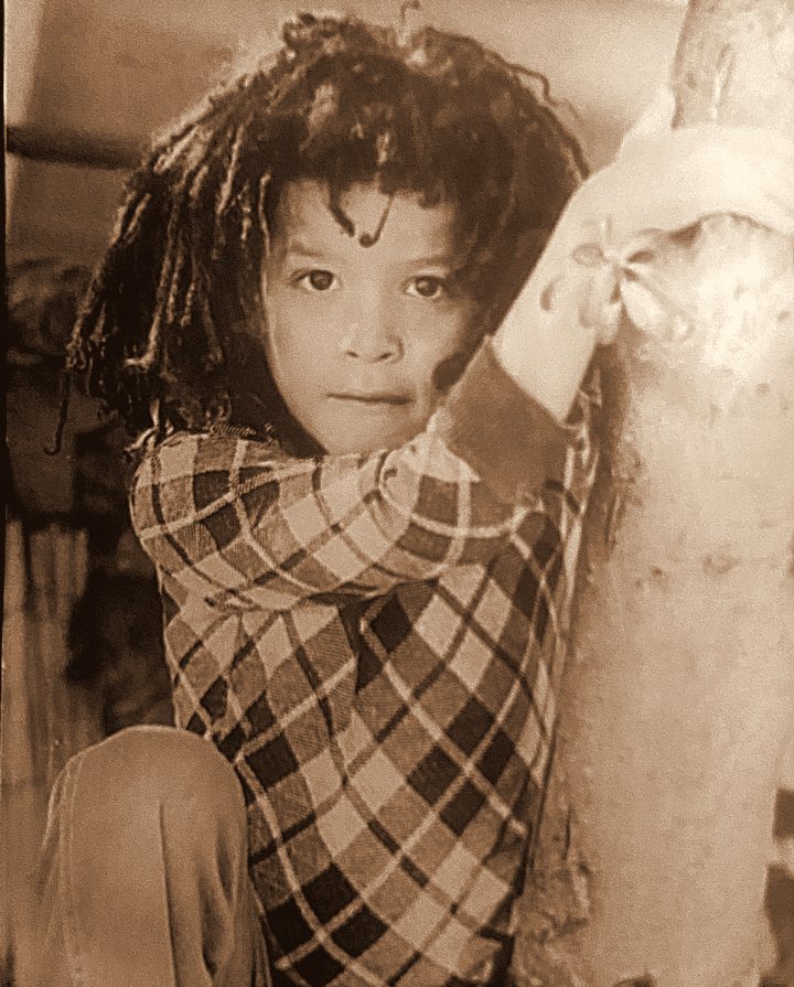 Raphael as a young child with long dreadlocks and a plaid shirt, holding onto a tree trunk and looking at the camera. A sepia photo.