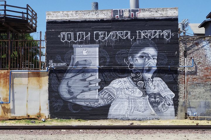 A large black-and-white mural on a brick wall beside railroad tracks. A woman in an embroidered blouse, with a bandana over her face, holds up a phone to take a selfie under the words South Central Dreams.