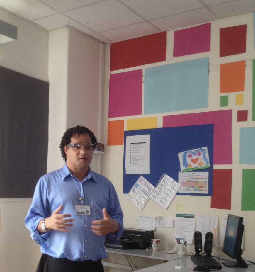 Raphael, wearing Google Glass, speaks to a class in front of a wall of bright paper squares.