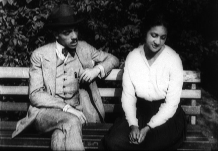 A man in a hat and suit sits on a park bench beside a young woman in a white blouse who looks down.